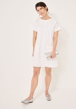 White short-sleeve dress with two front pockets, featuring a straight cut. Paired with silver flat shoes and holding a metallic clutch.