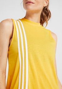 Top sin mangas amarillo con un cuello alto y tres rayas blancas en el hombro izquierdo. Fabricado con un tejido suave y ligero.