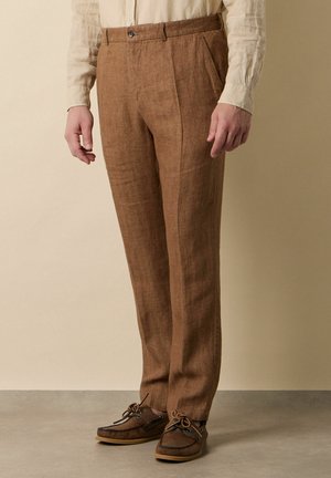 Man draagt bruine op maat gemaakte linnen broek, beige overhemd met knopen en bruine leren veterloafer, staand tegen een neutrale achtergrond.