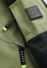 Olivengrøn jakke med sorte accenter, der har et tekstureret sort logo-patch og dekorative lynlåse med "BAKER" tekst i hvid.