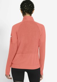 Chaqueta deportiva de coral claro con cuello alto, tejido texturizado y detalles perforados en las mangas y la espalda para mayor transpirabilidad.