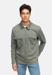 Olivgrüne Zip-Jacke mit Kragen, texturiertem Stoff und zwei großen Brusttaschen. Getragen über einem weißen Hemd, kombiniert mit blauen Jeans.