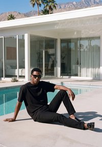 Uomo con abbigliamento nero e occhiali da sole seduto accanto a una piscina davanti a una casa moderna con porte di vetro e montagne sullo sfondo.