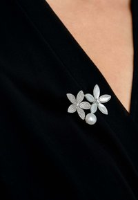 Silver floral brooch with one flower featuring clear gemstones and the other with a pearl center, set against a black fabric background.