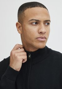 Svart zip-up genser med ribbestrikket krage og mansjetter, med en glidelås som har en stofftrekk. Modell som holder kragen foran en nøytral bakgrunn.
