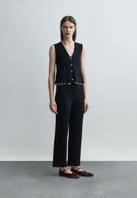 Kvinne iført svart ermeløs knappet topp og matchende bukser med små hengende anheng, kombinert med mørkerøde Mary Jane-sko.