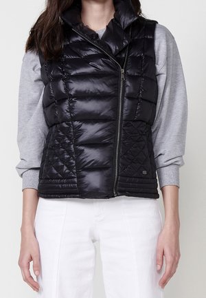 Frau trägt eine schwarze gesteppte Steppweste über einem grauen Langarmshirt, kombiniert mit weißen, hoch taillierten Hosen, steht vor einem einfarbigen Hintergrund.