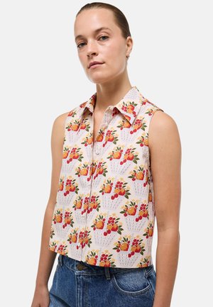 Femme portant une chemise sans manches à col avec un imprimé coloré de panier de fruits associée à un jean bleu en denim sur fond uni.
