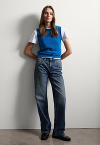 Jersey sin mangas azul sobre una camiseta blanca, combinado con vaqueros de denim azul de corte holgado y zapatos negros, frente a una pared sencilla.