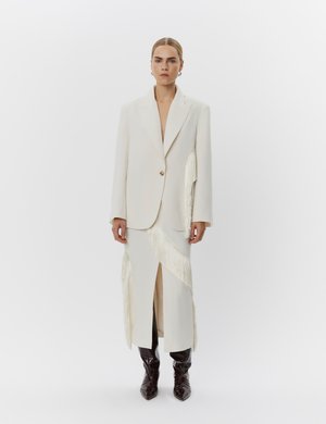 Vrouw die een offwhite op maat gemaakte blazer en bijpassende rok met franjes en een voorsplit draagt, gecombineerd met zwarte puntige laarzen.