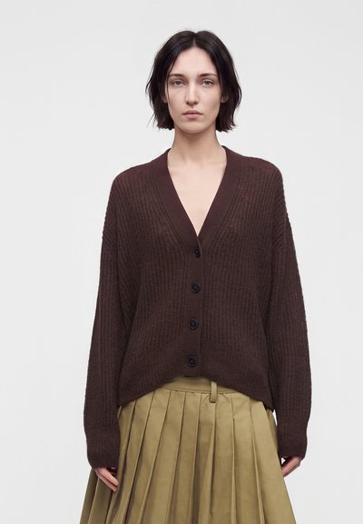 Femme portant un cardigan côtelé marron foncé boutonné et une jupe plissée vert olive, debout devant un fond clair uni.