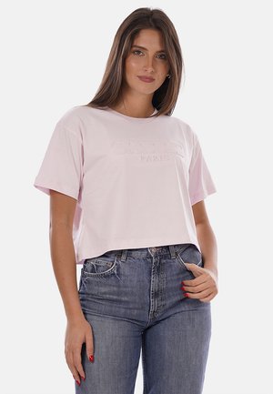 Mujer que lleva una camiseta corta rosa claro con texto en relieve y vaqueros azules de talle alto, de pie con una mano en el bolsillo.