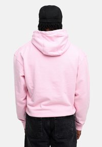 Short pink hoodie with a kangaroo pocket, adjustable hood, and ribbed cuffs, paired with black denim pants, view from the back.