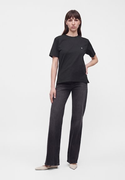 Femme debout portant un t-shirt noir à manches courtes, un jean droit foncé et des ballerines pointues beiges sur un fond clair uni.