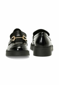 Mocassins en cuir verni noir avec un bout rond, présentant un détail de boucle en ton doré et une épaisse semelle en caoutchouc noir. Texture lisse sur toute la surface.