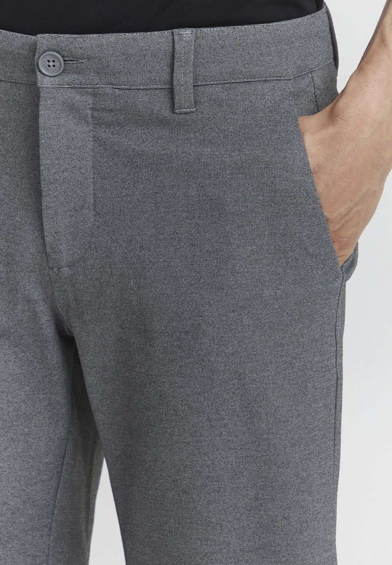 Gros plan sur un pantalon gris texturé avec une ceinture boutonnée et une main partiellement dans la poche avant.
