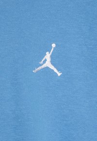 Un tessuto di cotone azzurro chiaro presenta un logo Jumpman ricamato in bianco, che rappresenta una figura in una posa di basket in volo.