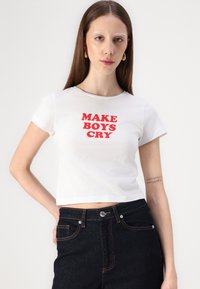 T-shirt branca corta com o texto vermelho "FAÇA OS RAPAZES CHORAR" centrado no peito. Modelo usa com jeans escuros de corte clássico.