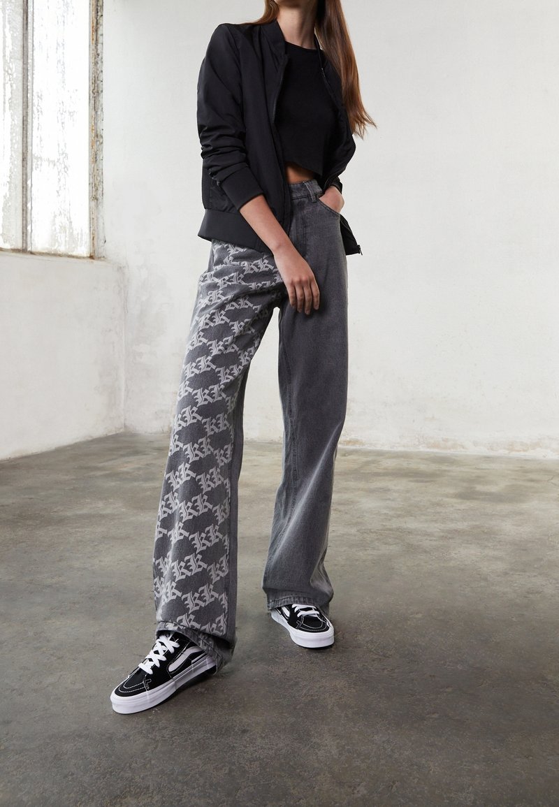 Personne portant un haut noir court, une veste noire, un jean ample gris avec un motif blanc sur une jambe, et des baskets noires, debout à l'intérieur.