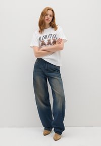 T-shirt grafico bianco con la scritta "MANHATTAN" e design di un orso abbinato a jeans blu larghi con accenti sbiaditi e scarpe beige.