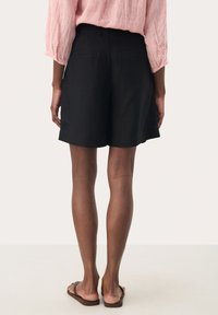 Zwarte linnen shorts met een losse pasvorm, voorzien van een middelhoge taille en twee achterzakken. Het model draagt bruine platte sandalen.