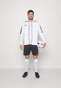 Witte sportjacket met zwarte accenten en logo, gecombineerd met zwarte korte broek en kniehoge witte sokken. Houdt een multi-kleurige voetbal vast.