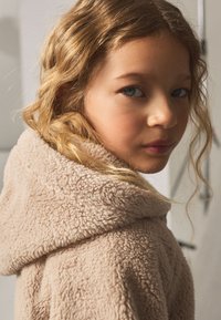 Jachetă bej, plușată, cu glugă și suprafață moale și texturată; purtată de o fată cu păr ondulat, de culoare castaniu deschis, prezentând un profil apropriat.