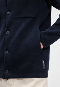 Ναυτικό πλεκτό σαλβάρι με υφασμάτινη υφή, διαθέτοντας κουμπιά, τσέπη στο στήθος και ετικέτα που λέει "Marco Polo", σε συνδυασμό με μπεζ παντελόνι.