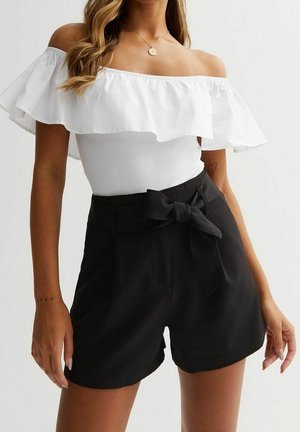 Frau trägt ein weißes, schulterfreies, volantes Oberteil und hochtaillierte schwarze Shorts mit einem vorderen Schleifengürtel, steht vor einem schlichten Hintergrund.