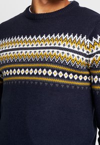Navy knitted sweater featuring a multicolored, geometric pattern in white, yellow, and gray on the chest. Crew neck design.