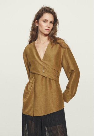 Blouse wrap verte olive à manches longues. Tissu doux et texturé, décolleté en V et détails drapés à la taille. Associée à une jupe plissée noire.