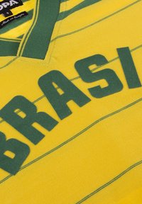 Gul sportströja med gröna ränder och stor text "BRAS". V-ringad design med mjuk textur. Etiketten anger "Tillverkad i Europa."