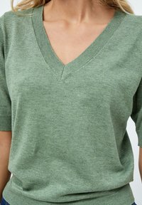 Zielony sweter z krótkim rękawem i dekoltem w kształcie litery V. Miękka, teksturowana tkanina o lekko luźnym kroju i prążkowanych krawędziach rękawów oraz dołu.