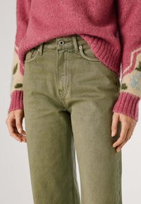 Los pantalones de algodón verdes presentan un corte recto con diseño de cinco bolsillos, combinados con un suéter de lana rosa con puños decorados.