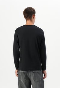 Person standing facing away wearing a black long-sleeve shirt and gray jeans with back pockets against a white background.