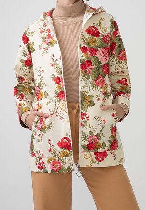 Beige gebloemde hoodie met rits, versierd met rode, roze en gele bloemen, gedragen over een beige geribbelde coltrui en beige broek, handen in de zakken.