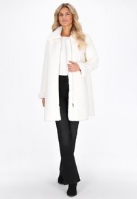 Vrouw in een witte mid-length jas met bontafwerking, witte top, zwarte flare jeans en zwarte laarzen, staand tegen een witte achtergrond.