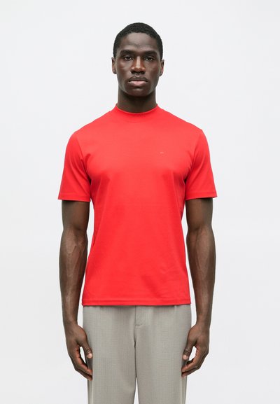 Homme portant une chemise rouge vif à manches courtes et un pantalon gris clair, se tenant devant un fond blanc uni.