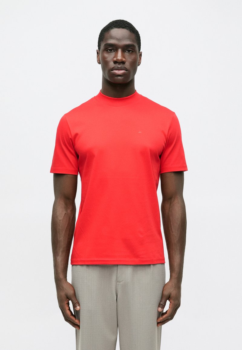 Άνδρας ντυμένος με ένα φωτεινό κόκκινο κοντομάνικο πουκ�άμισο και ανοιχτό γκρι παντελόνι, στέκεται μπροστά σε ένα απλό λευκό φόντο.