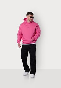 Sweatshirt à capuche rose avec une poche avant, pantalon noir et baskets blanches. Lunettes de soleil portées ; coupe décontractée avec des poignets et un ourlet côtelés.