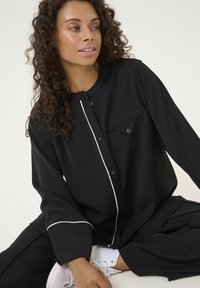 Melnā krekla ar apkakli, ar krēmkrāsas izliekumu gar priekšpusi un krūšu kabatu. Valkāts ar melnām biksēm un baltām kedām.