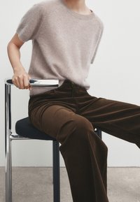 Un pull en cachemire beige clair à manches courtes associé à un pantalon en velours côtelé marron foncé. Une chaise métallique avec une main reposant sur son accoudoir.
