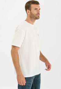 Witte katoenen t-shirt met korte mouwen, rechte zoom en een ronde halslijn. Heeft een klein grafisch ontwerp op de borst.