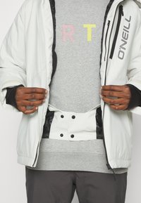 Witte geïsoleerde jas met zwarte accenten en rits, gedragen over een grijze sweatshirt met pastel geborduurde letters "R T."