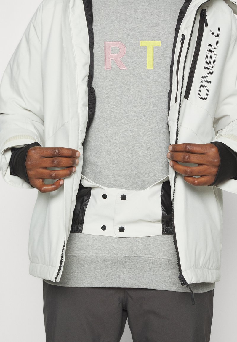 Weiße isolierte Jacke mit schwarzen Akzenten und Reißverschluss, getragen über einem grauen Sweatshirt mit pastellfarbenen gestickten Buchstaben "R T."