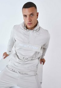 Ljusgrå atletisk outfit med en dragkedja och justerbar huva, med en framficka där "PROJECT X PARIS" står i vit text. Slät textur.
