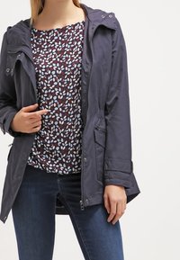 Navy waterproof jacket with a zip closure, adjustable hood, and side pockets, paired with a patterned top featuring blue and maroon leaves.