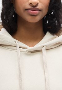 Μπεζ φούτερ με κουκούλα από υφή υφάσματος, με κορδόνια στο ντεκολτέ και άνετη εφαρμογή. Ομαλή συνολικ ή εμφάνιση.