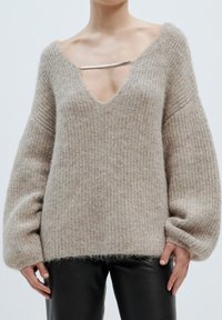 Hellbeige, oversized Pullover mit tiefem V-Ausschnitt, gerippter Textur und weiten Ärmeln. Mit einem silbernen Hardware-Akzent am Ausschnitt.