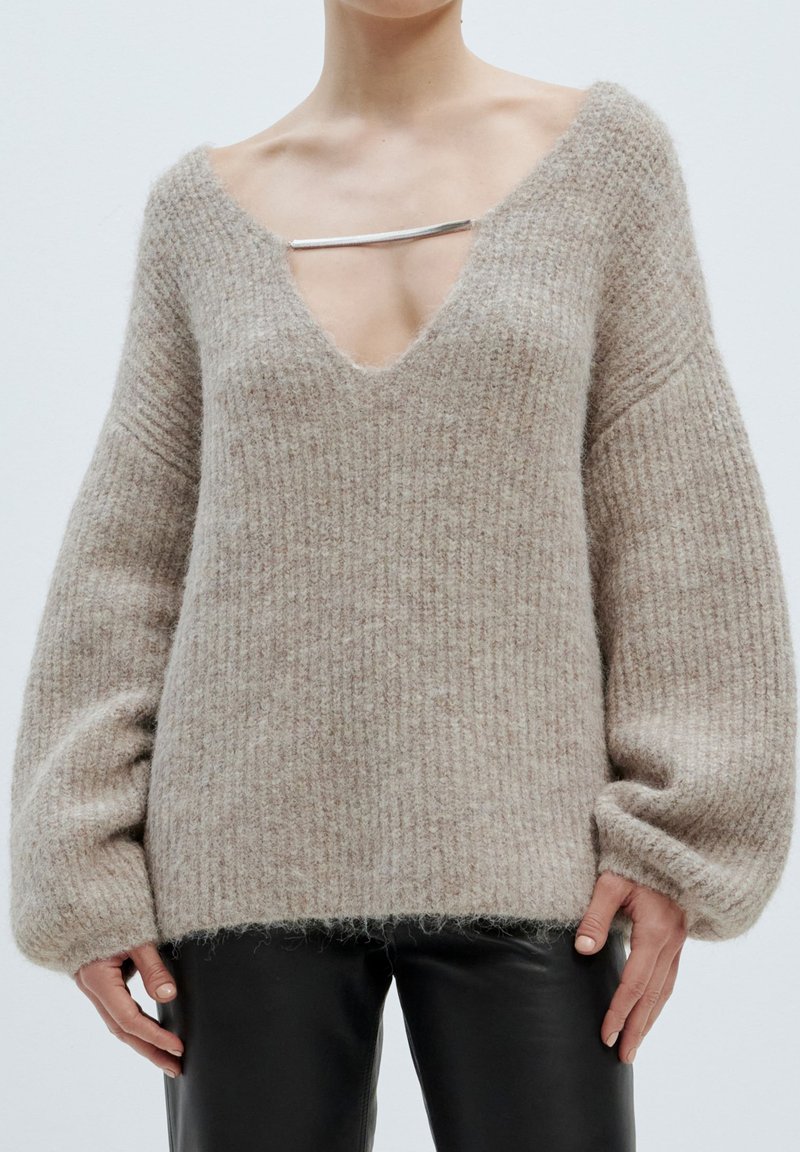 Hellbeige, oversized Pullover mit tiefem V-Ausschnitt, gerippter Textur und weiten Ärmeln. Mit einem silbernen Hardware-Akzent am Ausschnitt.
